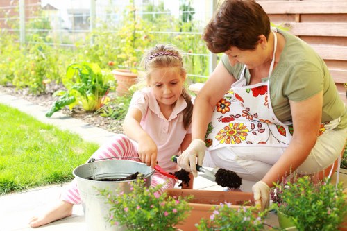 In a well-maintained garden with a lush green lawn, a woman and a young girl are engaged in gardening activities on a sunny day, sitting on the patio near a wooden fence. The woman, wearing a floral apron and light-colored clothing, is handling dark soil with gardening gloves, possibly planting or transferring plants into a rectangular garden bed edged with bricks. The girl, dressed in striped pink and white clothing, is assisting by holding a small gardening tool, showing curiosity and involvement. Around them are various garden elements including flower pots, potted plants with green foliage and purple flowers, and garden tools placed nearby. The background features a vibrant vegetable patch with leafy greens and herbs, framed by a trellis or wire fencing, indicating a thriving outdoor space designed for gardening. The overall environment is bright and cheerful, with natural sunlight illuminating the scene, reflecting active outdoor garden maintenance, typical of residential gardening services in the Pinner area.