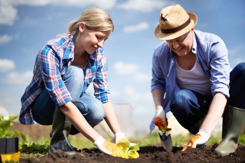 The image depicts a young woman and an older woman engaging in gardening outdoors in a well-maintained garden area, possibly in Pinner, with a bright blue sky and some fluffy clouds overhead. The younger woman, dressed in a plaid shirt and jeans, is kneeling on the soil, planting or tending to a young plant with green leaves, while the older woman, wearing a straw hat, light blue shirt, and gloves, is also kneeling and working with gardening tools. The garden features dark, freshly turned soil with some small gardening containers or pots nearby. In the background, there are lush green shrubs and a neatly trimmed hedge, indicating a landscaped front or back garden designed for outdoor enjoyment. The natural sunlight highlights the vibrant green foliage and the earthy tones of the soil, creating a warm and inviting atmosphere for outdoor maintenance, lawn care, and garden revitalisation, which is relevant to services offered by Landscapers Pinner.