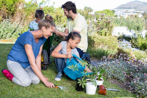 A family of four enjoying gardening in a well-maintained front yard garden scene, with a lush green lawn in the foreground and flower beds filled with blooming plants and shrubs. The mother is kneeling on the grass, planting or tending to a small shrub with gardening tools and a watering can nearby. A young girl is sitting on the grass, holding a blue bag with gardening gloves, while the father and a boy stand in the background, engaged in gardening activities amidst an array of colourful flowers and greenery. The garden features a mix of flowering plants, bushes, and a tidy lawn with visible soil patches, bordered by a variety of garden beds and ornamental plants. In the background, there are distant views of trees and buildings, suggesting a suburban setting near Pinner. The scene is lit with natural sunlight, indicating a bright, clear day, and overall conveys a peaceful outdoor environment suitable for gardening and outdoor maintenance.