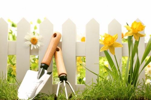 This image depicts a well-maintained garden scene featuring a white picket fence in the background with various plants and flowers growing in front of it. In the foreground, there is lush, green grass with a slight natural unevenness. To the right, a cluster of bright yellow daffodils with long green stems and leaves add vibrant colour to the scene. On the left side, a white daisy with a yellow centre is positioned near the fence among green foliage. Resting on the grass are gardening tools, including a trowel with a wooden handle and a garden fork with a similar handle, suggesting gardening or lawn care activities. The lighting is natural, indicating a sunny day, and the overall environment appears to be a neat and inviting backyard or front garden space, possibly in an area serviced by Landscapers Pinner, focusing on outdoor gardening and landscaping maintenance.
