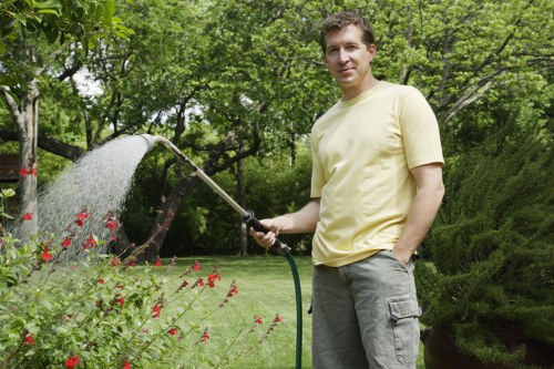 A man standing in a lush garden holding a garden hose, watering flowering plants with red blossoms. The garden features a well-maintained green lawn, bordered by various shrubs and hedges, with mature trees providing shade in the background. Paved paths and dense greenery create a natural indoor environment, and the scene is bathed in natural daylight, suggesting pleasant weather. The garden is part of an outdoor space suitable for gardening and landscaping maintenance, reflecting a professionally maintained landscape typical of residential gardens in Pinner, with visible attention to plant health and overall design—services offered by Landscapers Pinner.