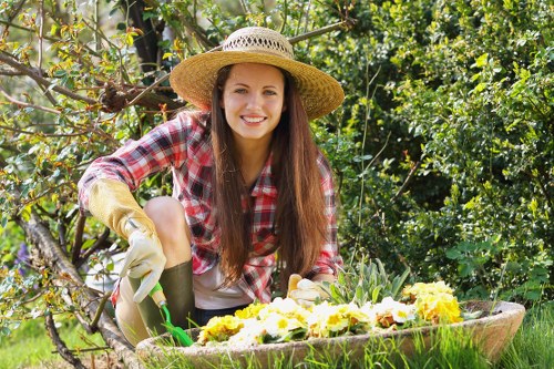 In this outdoor garden scene, a woman with long brown hair and a friendly smile is engaged in gardening activities in a lush, green environment that features a variety of plant life. She is wearing a wide-brimmed straw hat to protect herself from the sun, a red and white checkered shirt, beige gardening gloves, and light-colored trousers. She appears to be tending to a flower bed or container filled with blooming yellow and white flowers. The garden includes a well-maintained area with dense foliage, small shrubs, and trees in the background, indicating a healthy, cultivated outdoor space. The natural light suggests a bright day, and the vibrant colors of the plants contrast against the rich brown soil and greenery surrounding her. This scene highlights typical gardening activities, such as planting or caring for flowers in a residential garden setting, which is consistent with lawn care and outdoor maintenance services offered by companies like Landscapers Pinner, focusing on creating vibrant, well-kept gardens in Pinner or nearby areas within the UK.