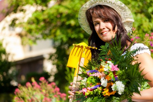 A woman smiling and holding a bouquet of colorful flowers and lush green foliage in a well-maintained garden with vibrant flowering plants in the background, a neatly trimmed hedge, and a paved patio area. She is wearing a wide-brimmed straw hat and casual clothing, standing in front of a lively garden landscape that includes a lawn, flower beds, and trees with fresh green leaves. The scene is outdoors on a bright, clear day, providing natural light that highlights the natural tones of the garden. This setting exemplifies a tidy and inviting outdoor space suitable for gardening and landscaping services offered by Landscapers Pinner, situated in or near Pinner, Middlesex, emphasizing professional garden transformation ideas and maintenance.