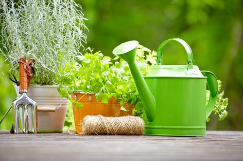 The image depicts a garden scene with a weathered wooden outdoor table displaying gardening tools and supplies. On the left, there is a small metal hand fork with a wooden handle, resting against a beige ceramic pot filled with tall, silver-green foliage. Next to this, there is a terracotta pot containing bright green, leafy herbs or plants, with vibrant foliage extending outward. To the right of the pots, a coiled jute twine lies on the table, suggesting gardening activity. Prominently positioned on the right side of the table is a bright green plastic watering can with a large, curved handle and a long, narrow spout. The background reveals a lush, green garden environment with blurred trees and foliage, indicating an outdoor space on a sunny day, ideal for gardening or outdoor maintenance activities. The scene emphasizes elements typical of gardening and outdoor care, aligning with services provided by Landscapers Pinner in the Pinner area, close to postcode HA5, creating a natural connection to professional gardening and landscaping services.