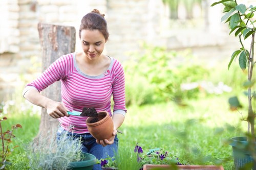 A young woman with dark hair tied back, wearing a pink and white striped long-sleeve top, is tending to a flower bed in a garden, using a small trowel to plant or weed in a terracotta pot. The garden features a well-maintained lawn with lush green grass, bordered by flower beds containing various plants, some with purple flowers and others with green foliage. In the background, there are trees, a stone wall, and a wooden garden bench, indicating a landscaped outdoor space with natural light and mild weather conditions. The scene reflects typical gardening activity, supported by local landscaping services such as those offered by Landscapers Pinner, with a focus on garden maintenance and outdoor aesthetics in a suburban area around Pinner, Middlesex.