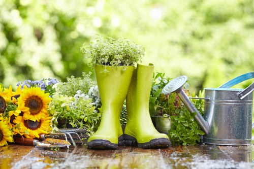 A garden scene featuring a weathered wooden outdoor table with a variety of gardening tools and decorative elements arranged on its surface. Prominently, there are a pair of bright green rubber boots filled with small green plants, serving as unique planters. To the right of the boots, a metallic watering can with a blue handle is positioned, alongside a matching spray nozzle resting on the table. On the left side, a bunch of vibrant yellow sunflowers and other mixed flowering plants add colour and natural textures to the setting. Behind the boots, there are potted plants with green foliage, suggesting a well-maintained garden environment. The background is softly blurred, revealing hints of lush greenery and trees under natural daylight conditions, indicating a mild, sunny outdoor day typical of gardens in Pinner, Middlesex. This scene, captured in natural light, reflects outdoor gardening activities and could be associated with services offered by Landscapers Pinner, specialising in garden design, planting, and outdoor space maintenance in the local area.