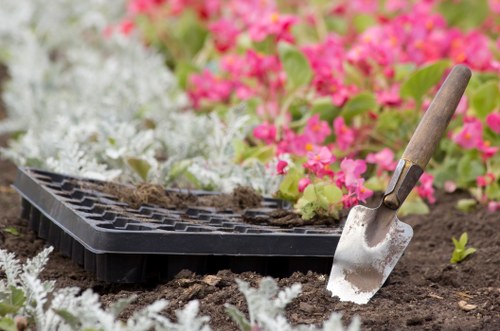 A gardening scene in a landscaped outdoor area, showing a close-up of dark soil being prepared for planting, with an empty black seed tray partially embedded in the earth. To the right, a small hand trowel with a wooden handle rests in the soil, indicating ongoing planting or soil cultivation. Surrounding the planting area, there are soft, white ground cover plants and vibrant pink flowering plants in the background, creating a colorful contrast with the natural tones of the soil and tools. The garden appears to be part of a neatly maintained front or back garden space, with a focus on flower bed preparation, and is typical of outdoor garden care services such as those provided by Landscapers Pinner, reflecting a well-kept, visually appealing outdoor environment, possibly in or near Pinner, Middlesex, where gardening and landscaping work is common. The scene is illuminated by natural daylight, suggesting a clear, mild-weather day suitable for gardening activities.