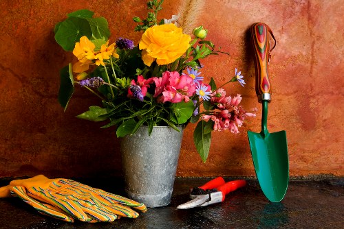 A close-up image of a gardener's work station featuring a large grey concrete pot filled with a vibrant assortment of flowers, including yellow, pink, purple, and white blooms, along with green foliage. To the right of the flower pot are small red gardening gloves, a pair of silver pruning shears, and a small green-handled trowel with a wooden handle, all resting on a dark garden surface. In the background, there is a warm-toned wall with a slightly textured surface, suggesting an outdoor or garden shed environment. The scene emphasizes gardening tools and flowering plants, indicating recent planting or garden maintenance activities, typical of professional gardening services in the Pinner area, UK. The overall composition highlights the tools and floral display that are likely used in outdoor garden care and planting tasks as offered by landscaperspinner.co.uk.