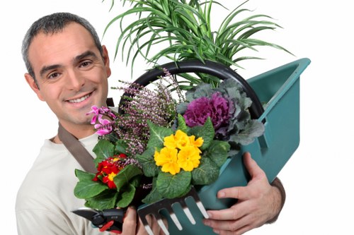 A man with short dark hair and a friendly smile is holding a black plastic garden container filled with a variety of flowering plants and green foliage. The plants include yellow, purple, and pink flowers, surrounded by broad green leaves. He is standing outdoors in a garden setting, with a wooden fence and some additional greenery visible in the background. The scene is lit with natural daylight, suggesting a bright, clear day. The man appears to be a gardener or landscaping professional, possibly associated with Landscapers Pinner, and the garden environment hints at professional gardening or landscaping services such as planting or garden maintenance. The container is placed in front of him, with some gardening tools or watering can also visible nearby, emphasizing outdoor gardening tasks. The overall scene highlights attention to detail in garden planting and care, suitable for a gardening service website aimed at enhancing outdoor spaces in the Pinner area, with lush natural elements and vibrant plant life presented in a professional manner.