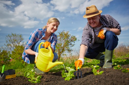 A woman and a man are working together in a garden during daylight hours, planting young leafy green plants in dark, well-tilled soil with small garden tools nearby. The woman is smiling and wearing a blue checked shirt, yellow gardening gloves, and jeans, while the man, dressed in a straw hat, a checked shirt, and rubber boots, is also smiling as he tends to the plants. They are in an outdoor garden space that features a grassy lawn and a hedge in the background, with a clear blue sky and some scattered clouds above. The scene conveys active garden maintenance, possibly part of a landscaping service by Landscapers Pinner, emphasizing healthy plant growth and outdoor environmental care in a pleasant, rural setting near Pinner, Middlesex.
