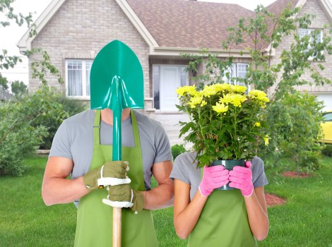 In a well-maintained front garden, two gardeners wearing grey T-shirts and bright green aprons are engaged in outdoor work. The gardener on the left is holding a large green garden spade with a shiny metal blade, wearing green gloves, but their face is hidden by a teal protective hood. The gardener on the right, also in gloves, is holding a potted plant with vibrant yellow flowers and green foliage close to their face, partially obscuring it. Behind them, the garden features a neatly trimmed lawn with lush, dense grass, bordered by small, young trees and shrubs with green leaves. The area includes a paved pathway or patio section, possibly made of concrete or stone, leading toward a beige brick house with white-framed windows and a grey roof, indicative of a suburban residential environment. The scene is lit by natural daylight, with clear weather conditions, contributing to a bright and inviting outdoor space. This setting exemplifies professional gardening activities with an emphasis on outdoor landscaping and plant care, suitable for a gardening service website focused on sustainable landscaping practices in Pinner, Middlesex, UK, supporting the themes of recycling and environmental responsibility promoted by Landscapers Pinner.