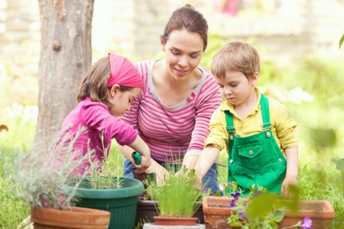 A woman with dark hair tied back, wearing a pink and white striped long-sleeve shirt, is gardening outdoors in a residential backyard garden. She is assisting two children, a girl in a purple jacket and pink headband, and a boy in a yellow shirt with green overalls, as they tend to potted plants on a garden table. The garden features a lush green lawn, a large tree trunk on the left, and various potted plants and flower beds with vibrant foliage. The background shows a fence and some flowering plants, indicating a well-maintained outdoor space during a sunny day, suitable for gardening activities. The scene reflects professional gardening support by Landscapers Pinner, emphasizing outdoor maintenance and lawn care within a family-friendly garden environment in Pinner, northwest London, UK, aligning with local gardening services and outdoor safety practices.